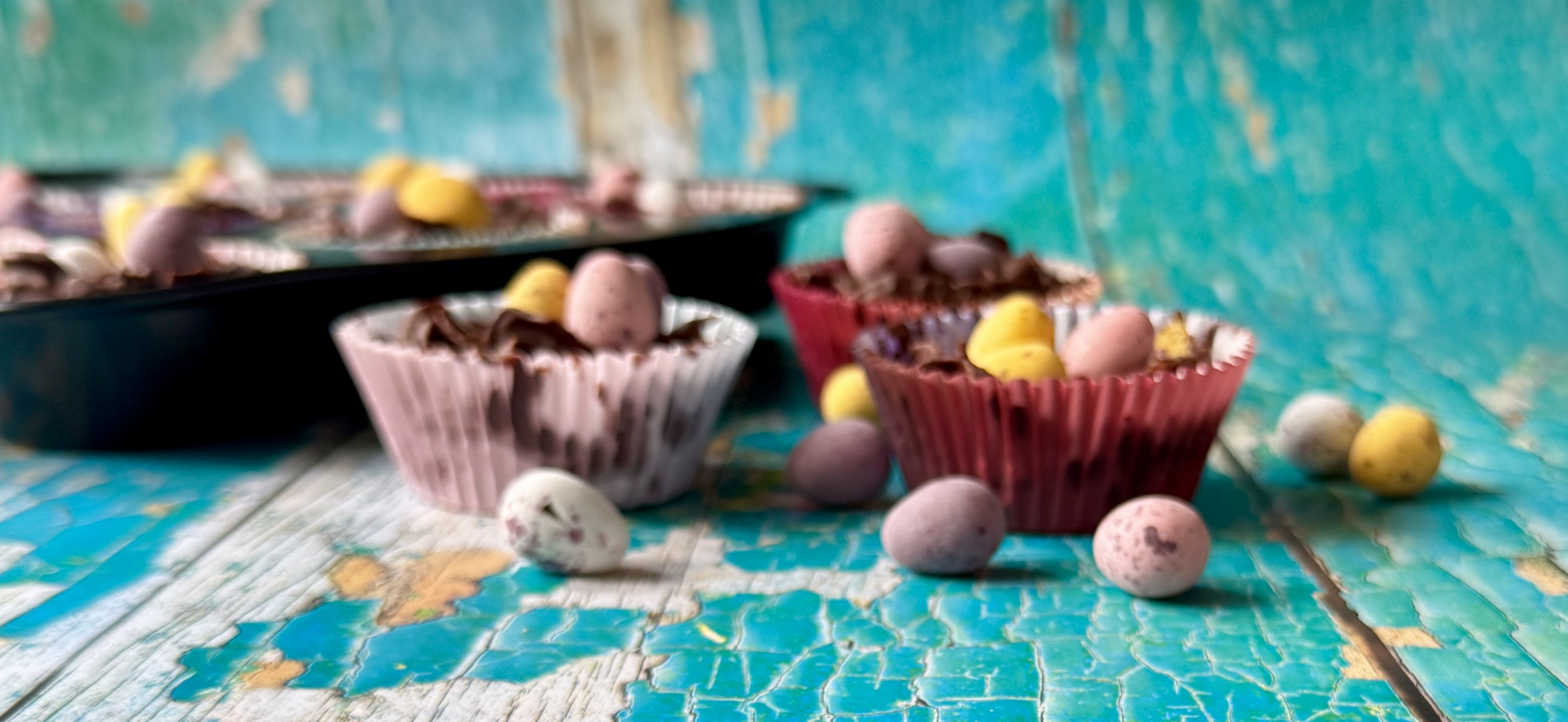 Close-up of Easter cornflake cakes in paper cases topped with Mini Eggs on a distressed turquoise wooden table, with a cupcake tray blurred in the background.